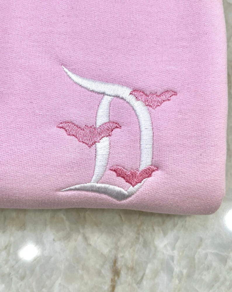 May include: A light pink sweatshirt with a white embroidered letter "D" and three pink bats. The bats are positioned around the letter. The sweatshirt is made of a soft, textured fabric.