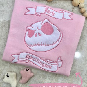 May include: A light pink sweater featuring an embroidered Jack Skellington design. The design includes the words "Jack" and "Skellington" on banners. Two small felt ghost figures are next to the sweater.