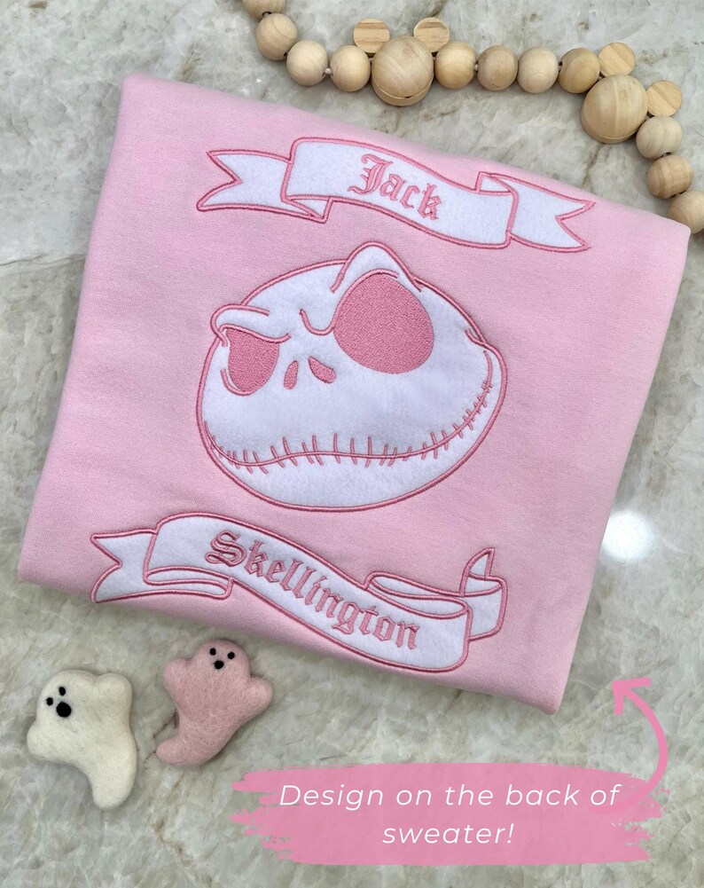 May include: A light pink sweater featuring an embroidered Jack Skellington design. The design includes the words "Jack" and "Skellington" on banners. Two small felt ghost figures are next to the sweater.