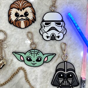 May include: Four embroidered keychains featuring Star Wars characters: a brown Chewbacca, a white Stormtrooper, a green Grogu, and a gray Darth Vader. Each keychain has a gold-toned metal lobster clasp and ring attached to the top of the character head.