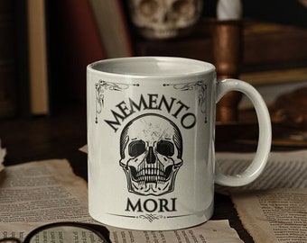 May include: White ceramic mug with a black skull and the text "Memento Mori" printed on it. The mug is sitting on a table with old books, a magnifying glass, and a skull figurine.