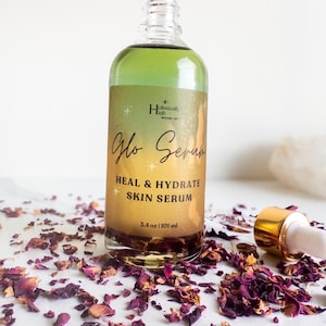May include: A glass bottle of "Glo Serum"  skin serum with a gold lid and label. The label reads "Holistically High" and "Heal & Hydrate Skin Serum". The bottle is sitting on a white surface with rose petals scattered around it.