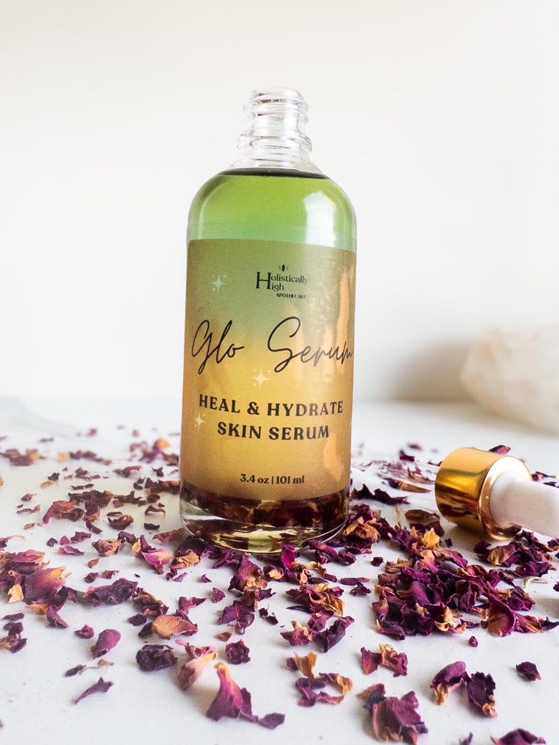 May include: A glass bottle of "Glo Serum"  skin serum with a gold lid and label. The label reads "Holistically High" and "Heal & Hydrate Skin Serum". The bottle is sitting on a white surface with rose petals scattered around it.
