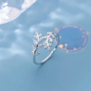 May include: A silver ring with a delicate design of two intertwined branches with small, clear stones. The ring is on a light blue background.