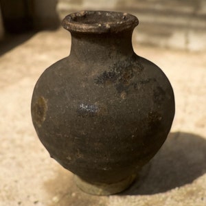 May include: A dark brown, round, ceramic pot with a wide mouth and a slightly flared rim. The pot has a smooth, matte finish and appears to be very old.