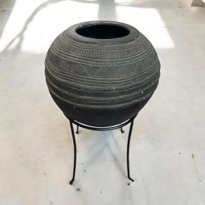May include: A large, round, black clay pot with intricate carvings sits on a black metal stand. The pot has a wide opening at the top and is decorated with a series of horizontal lines and geometric patterns.