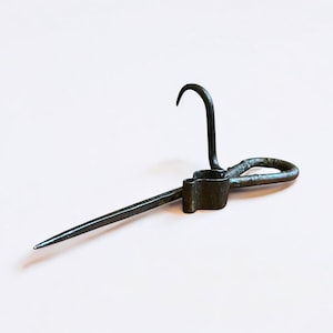 May include: A dark metal tool with a hook, a pointed end, and a looped handle. The tool has a rectangular section with a small opening. The hook curves downward, and the pointed end extends outward.