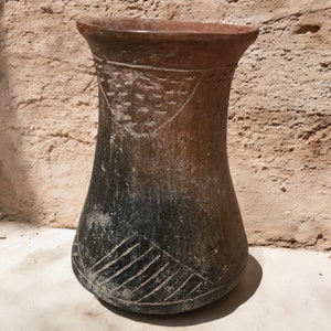 May include: A handmade, brown ceramic vase with a wide base and flared rim. The vase features etched geometric patterns and a dark, ombre effect from the base to the middle. The top rim has a series of small, carved holes.