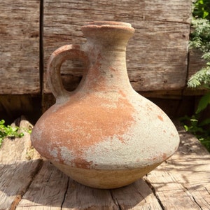 May include: A rustic terracotta pitcher with a handle, showing signs of age and wear. The pitcher has a light brown color with a white, weathered patina.