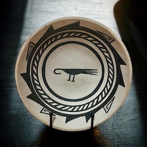 May include: A cream-colored ceramic bowl with a black geometric design. The bowl features a central bird illustration and a repeating pattern around the rim. The bowl is displayed on a black metal stand.