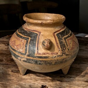 May include: A small, three-legged ceramic pot with a rounded body and a wide opening. The pot is decorated with black and red geometric patterns on a tan background.