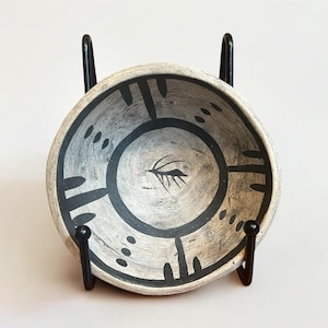 May include: Handmade ceramic bowl with a gray and white speckled finish. The bowl features a black circular design with a central leaf-like motif and black geometric patterns. The bowl is displayed on a black metal stand.