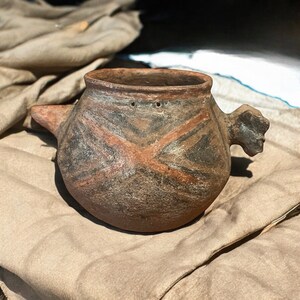 May include: A hand-painted, earthenware vessel with a spout and a handle. The pot features a rounded body with a dark brown base and a reddish-brown and black geometric design. The handle is shaped like a stylized human head.