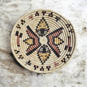 May include: A round, woven basket with a geometric design. The basket features concentric circles of natural beige, black, pink, and yellow fibers. The design radiates from the center, creating a symmetrical pattern. The basket is likely handmade.