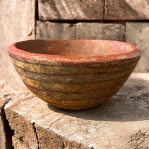 May include: A small, round, terracotta bowl with a red rim and brown and yellow stripes. The bowl is sitting on a rough, brown surface.