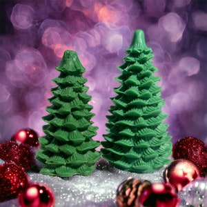Op de afbeelding: Twee groene kerstboomkaarsen op een glinsterend zilveren oppervlak omringd door rode en gouden kerstballen.