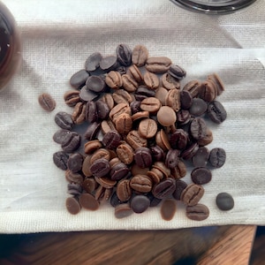 May include: A close-up of a pile of dark and light brown chocolate chips on a white fabric surface. The chips are arranged in a random pattern and some are slightly overlapping.