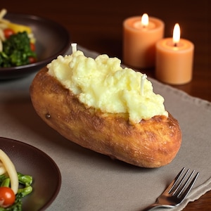 May include: A unique candle shaped like a baked potato, topped with a creamy, yellow wax resembling mashed potatoes. The candle sits on a gray placemat, with two lit orange candles in the background.