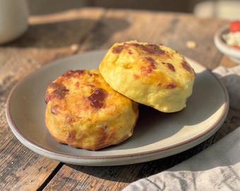 May include: Two round, golden-brown baked goods with visible melted cheese patches, stacked slightly on a light-colored ceramic plate. The surface of the items appears textured and uneven, showing a mix of pale yellow and toasted brown colors.