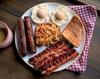 May include: A white ceramic plate featuring a breakfast meal consisting of two fried eggs with yellow yolks, two browned sausages, a golden-brown hash brown patty, a slice of cooked ham, and three strips of crispy bacon arranged on the plate.