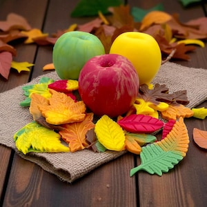 Pode incluir: Uma natureza morta com três maçãs verdes, vermelhas e amarelas, rodeadas por decorações em forma de folhas coloridas. As folhas são em tons de vermelho, laranja, amarelo e castanho, dispostas sobre um tecido de juta e uma superfície de madeira.