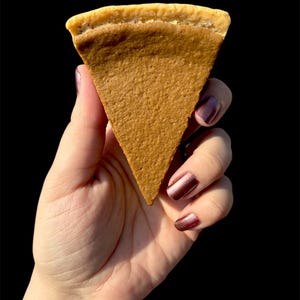 May include: A hand holding a triangular slice of pie against a black background. The pie has a light brown filling and a golden-brown crust. The hand has polished nails. The pie slice is a dessert.