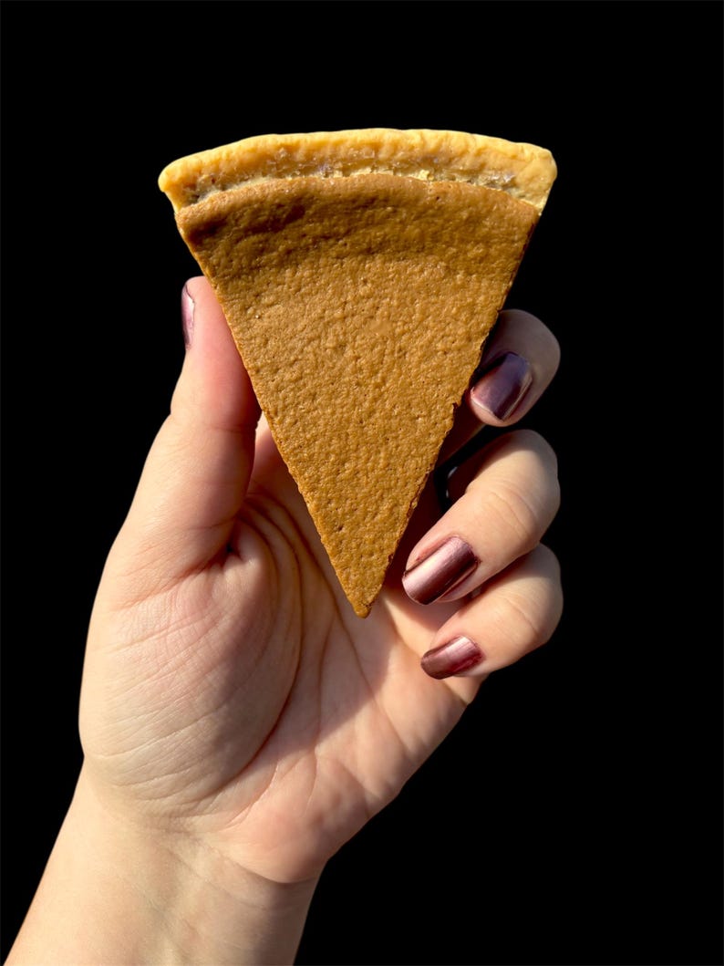 May include: A hand holding a triangular slice of pie against a black background. The pie has a light brown filling and a golden-brown crust. The hand has polished nails. The pie slice is a dessert.