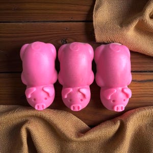 Könnte beinhalten: Drei rosa Seifen in Schweineform liegen auf einer Holzoberfläche, mit einem beigen Tuch im Hintergrund. Die Seifen haben detaillierte Schnauzen, Ohren und einen Ringelschwanz. Die Seifen haben eine leuchtende, einfarbige rosa Farbe.