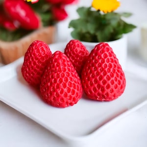 Peut inclure: Quatre bougies en forme de fraise rouge sur une assiette carrée blanche. Les bougies sont disposées en demi-cercle.