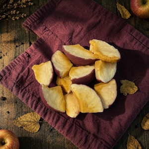 Peut inclure: Un groupe de morceaux de pommes tranchés avec une chair jaune pâle et une peau rouge foncé, disposés sur un tissu plié bordeaux profond. Les tranches de fruits sont coupées en quartiers, montrant une texture ferme et une couleur naturelle.