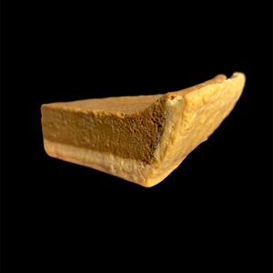 May include: A single slice of pie with a light brown top layer, a darker brown middle layer, and a light-colored bottom layer. The pie slice has a slightly uneven shape and is set against a black background.
