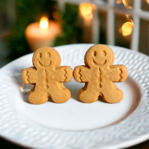 Puede incluir: Dos galletas de hombre de jengibre con caras sonrientes en un plato blanco. Las galletas son de color marr&oacute;n claro y tienen una superficie ligeramente texturizada.