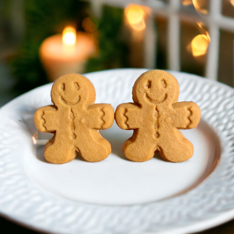 Puede incluir: Dos galletas de hombre de jengibre con caras sonrientes en un plato blanco. Las galletas son de color marr&oacute;n claro y tienen una superficie ligeramente texturizada.