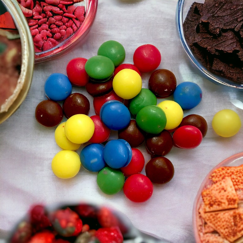 May include: A variety of colorful chocolate candies in red, yellow, green, blue, and brown. The candies are arranged in a pile on a white surface.