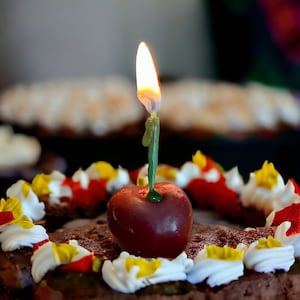 Puede incluir: Un pastel de chocolate con glaseado blanco y rojo y una sola vela encendida en el centro. La vela es verde y tiene una cereza roja en la parte superior.