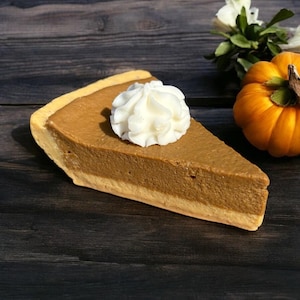 May include: A slice of pumpkin pie with a dollop of whipped cream. The pie has a golden-brown crust and a rich, orange filling. A small, orange pumpkin and white flowers are in the background. The pie is on a dark wooden surface.