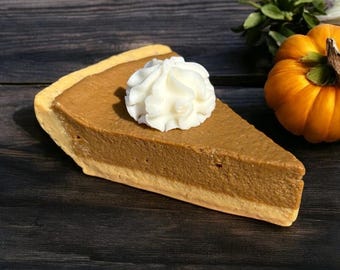May include: A slice of pumpkin pie with a dollop of whipped cream. The pie has a golden-brown crust and a rich, orange filling. A small, orange pumpkin and white flowers are in the background. The pie is on a dark wooden surface.
