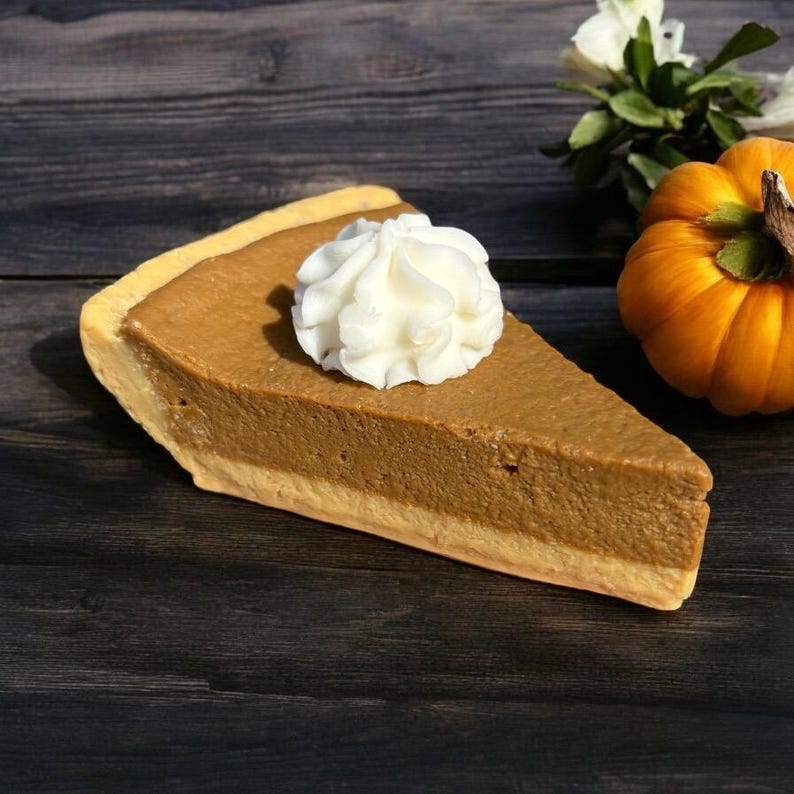 May include: A slice of pumpkin pie with a dollop of whipped cream. The pie has a golden-brown crust and a rich, orange filling. A small, orange pumpkin and white flowers are in the background. The pie is on a dark wooden surface.