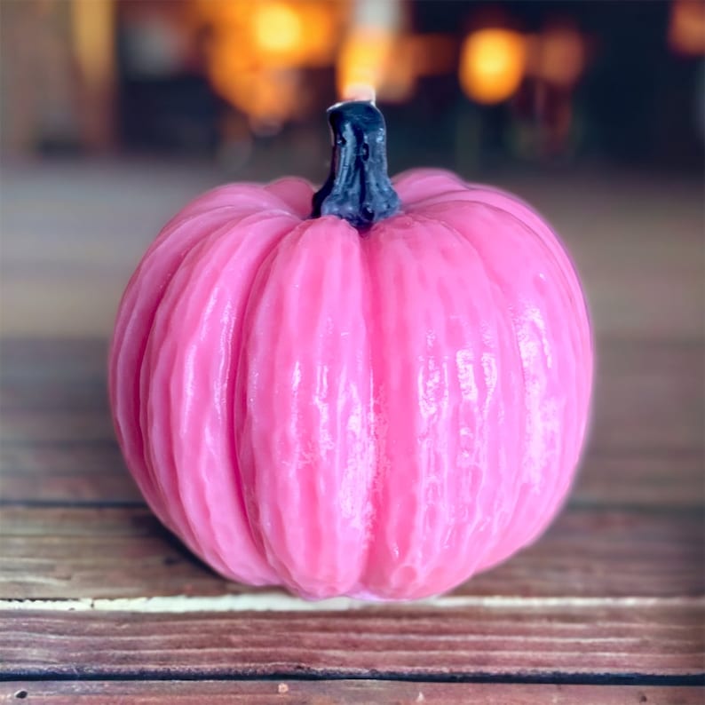 May include: A pink pumpkin-shaped candle with a black stem. The candle has a textured surface and is perfect for fall decor.