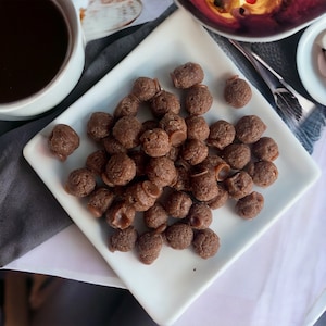 May include: A white plate filled with chocolate-covered cereal balls. The cereal balls are round and have a slightly irregular shape. The plate is on a white surface.