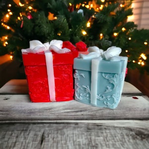 May include: Two decorative gift boxes, one red with white ribbon and one blue with white ribbon, both with ornate designs. The boxes are sitting on a wooden surface in front of a Christmas tree with lights.