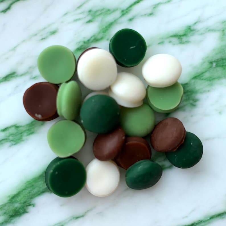 May include: A collection of round wax melts in various colors, including green, brown, and white. The wax melts are arranged in a pile on a marble surface.