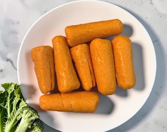 May include: A white plate holds seven cylindrical orange food items with a smooth texture. Beside the plate, a cluster of fresh green broccoli florets with thick stalks is visible, showing detailed leaves and stems on a light gray surface.