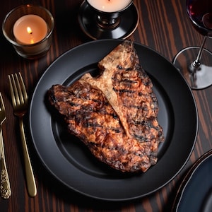 Puede incluir: Un filete T-bone a la parrilla en un plato negro, con marcas de parrilla. El filete está rodeado por un tenedor dorado, una vela encendida en un portavelas de vidrio y una copa de vino tinto. La mesa es de madera oscura.