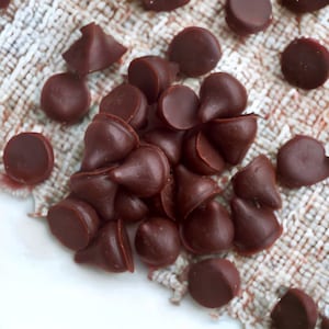 May include: A close-up of a pile of dark brown chocolate chips on a white surface with a textured fabric background.
