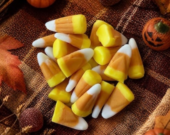 May include: A pile of triangular candy corn pieces, each featuring a white tip, an orange center, and a yellow base. The candies are arranged in a cluster, surrounded by small decorative pumpkins and textured autumn-colored fabric elements.