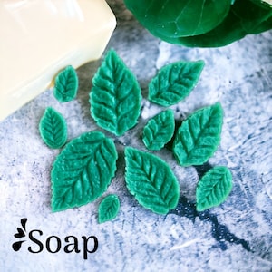Könnte beinhalten: Ein Set mit 10 grünen, blattartigen Silikonformen zum Seifenherstellen. Die Formen sind auf einer grauen, strukturierten Oberfläche angeordnet. Das Wort "Soap" ist in Schwarz in der linken unteren Ecke des Bildes geschrieben.