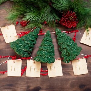 May include: Three green Christmas tree-shaped candles on a wooden surface with red and white string lights and small paper tags with the word "Elf" written on them.