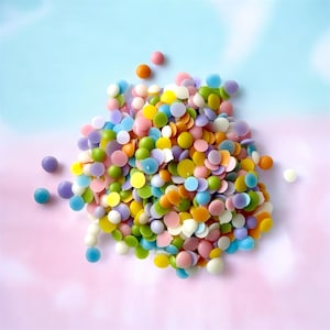 Puede incluir: Un montón grande de confeti redondo de colores, en tonos de rosa, morado, azul, verde, amarillo y naranja. El confeti está esparcido sobre un fondo azul claro y rosa.