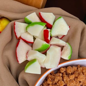 May include: A close-up of white, red, and green candy apple slices. The candy pieces are arranged on a tan cloth, with a bowl of brown crumble in the bottom right corner. A yellow and red apple is visible in the background.